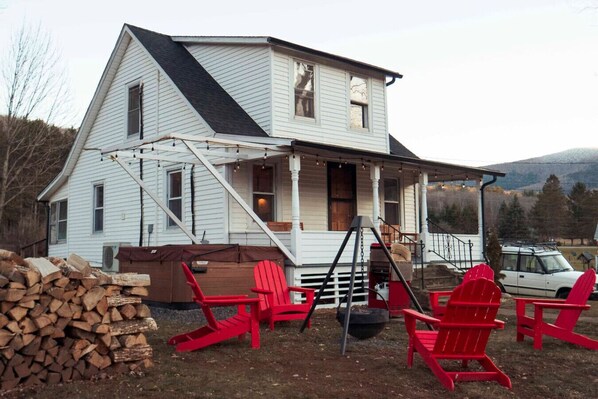 A two-story white house with a porch, a firewood stack, and red Adirondack chairs. - Charming Mtn Stay | Views, Hot Tub, Prime Location (West Kill)