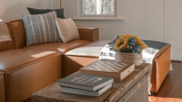 Een gezellige woonkamer met een leren bank, een houten salontafel met boeken, een vloerlamp en een raam met uitzicht op bomen.