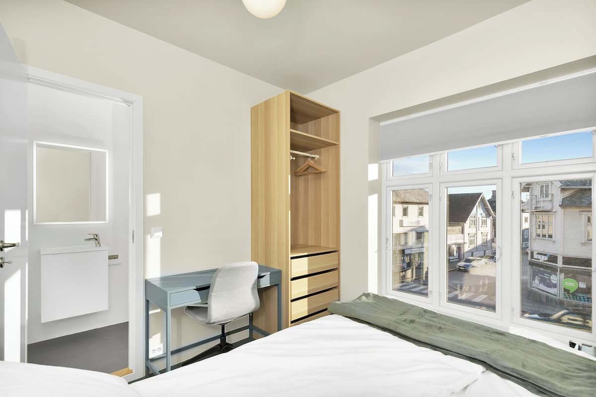 Un dormitorio con cama, escritorio con silla, armario y ventana con vista a edificios.