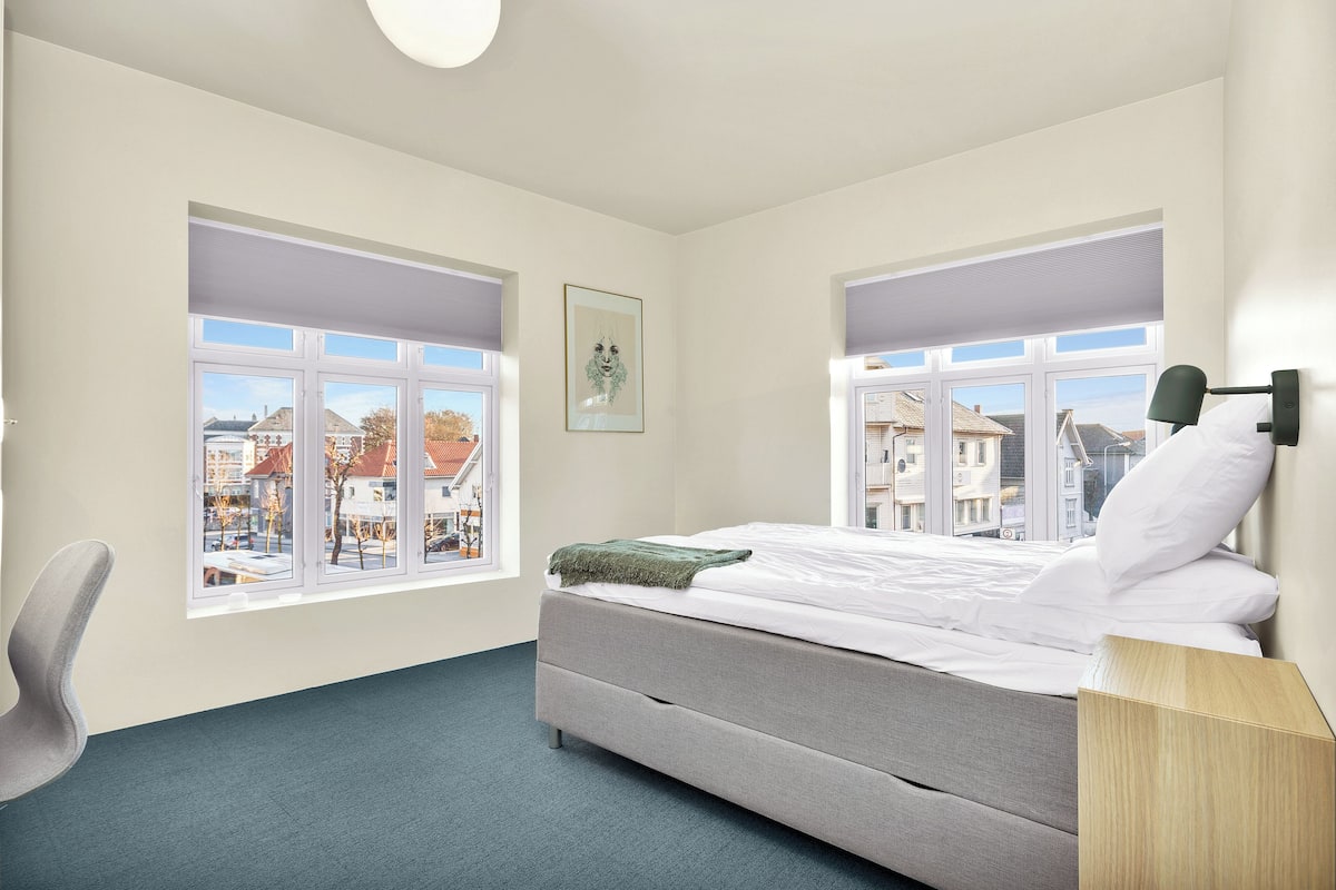 Un dormitorio moderno con una cama, dos ventanas, una silla y una mesita de noche.
