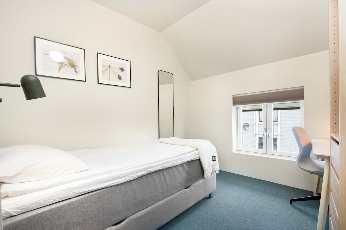 Un dormitorio con cama, escritorio, silla, ventana y dos cuadros en la pared.