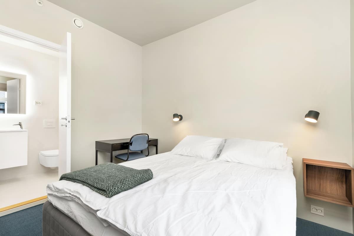 Un dormitorio con cama, escritorio, silla, mesita de noche y lámpara de pared.