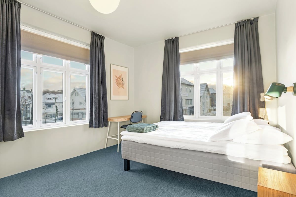 Habitación de hotel con cama, escritorio y silla. Hay ventanas con cortinas, un cuadro en la pared y una lámpara en la mesita de noche.