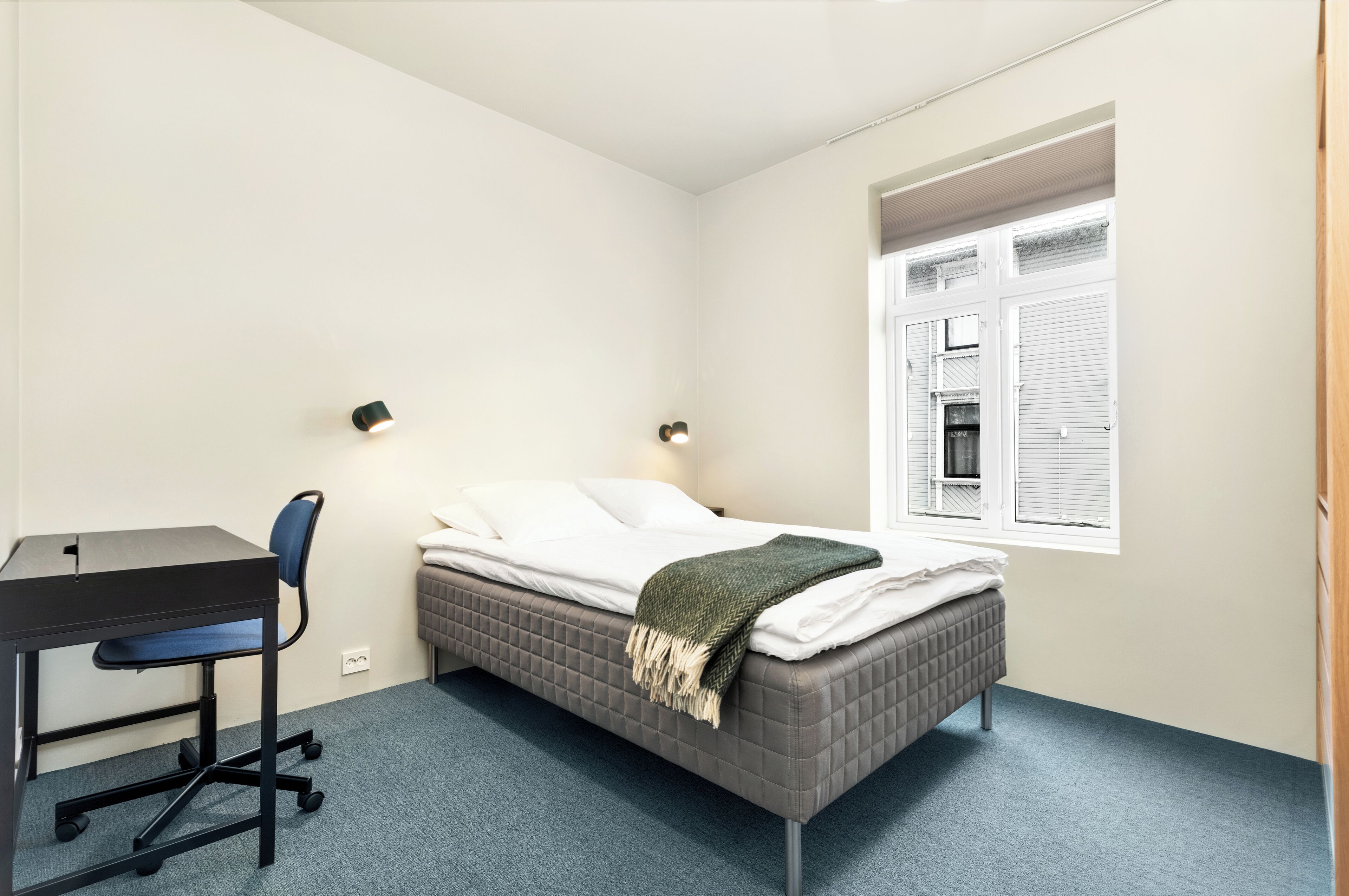 A bedroom with a bed, desk, chair, and a window with blinds.