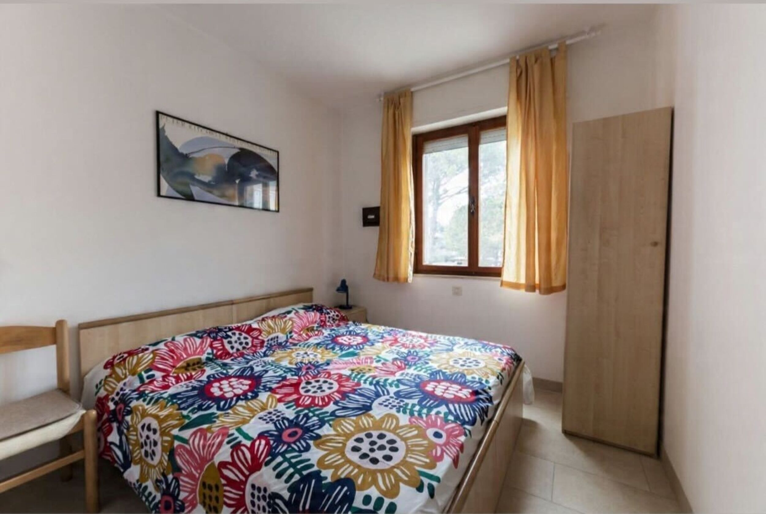 A bedroom with a bed, a nightstand, a window with curtains, a wardrobe, and a painting on the wall.