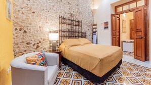 A bedroom with a stone wall, a bed with a yellow bedspread, a white chair, and a patterned floor. - CASA LA ESCONDIDA (Mérida)