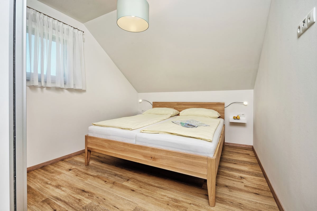 A bedroom with a wooden bed, white curtains, a ceiling light, and a small shelf with a fruit basket.