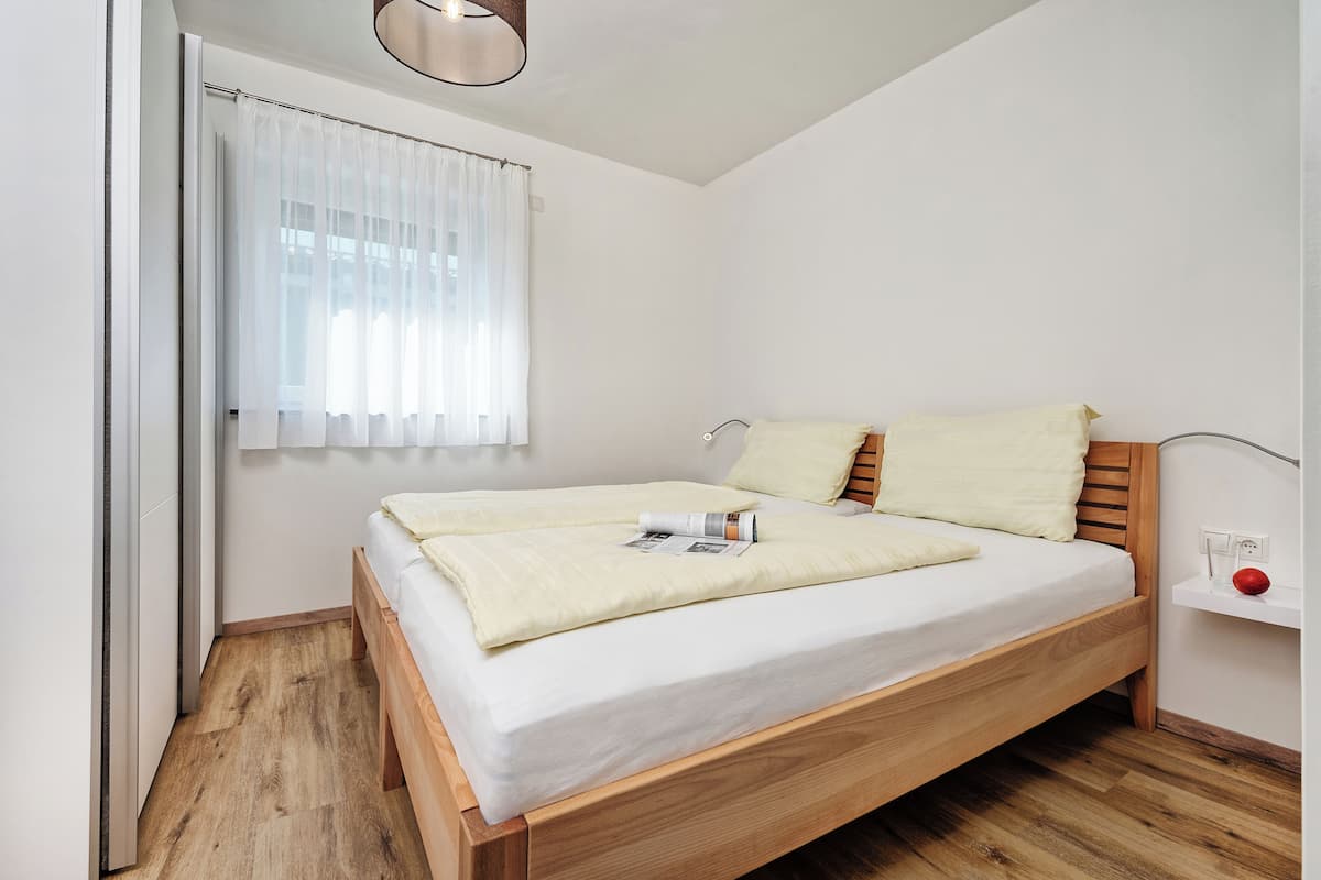 A single bed with a wooden frame and white bedding, a bedside table with a red object, and a window with sheer curtains.