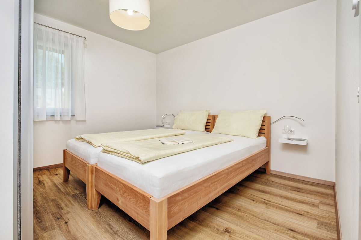 A bedroom with a wooden bed, white walls, a window with sheer curtains, and a bedside table with a lamp.