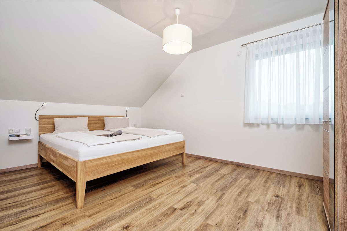 A bedroom with a wooden bed, a bedside table, a ceiling light, and a window with sheer curtains.