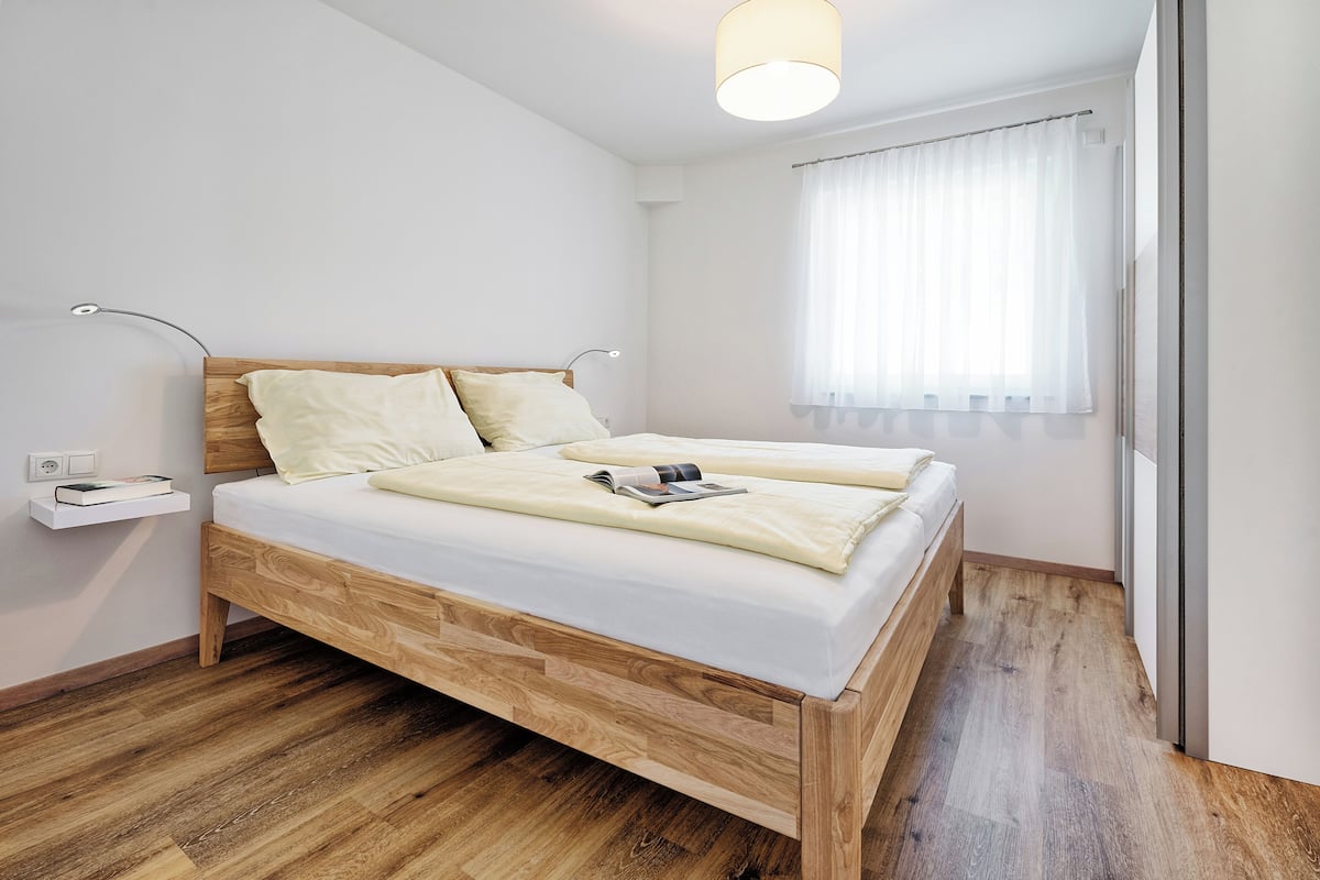 A modern bedroom with a wooden bed, a bedside table with a lamp, and a window with sheer curtains.