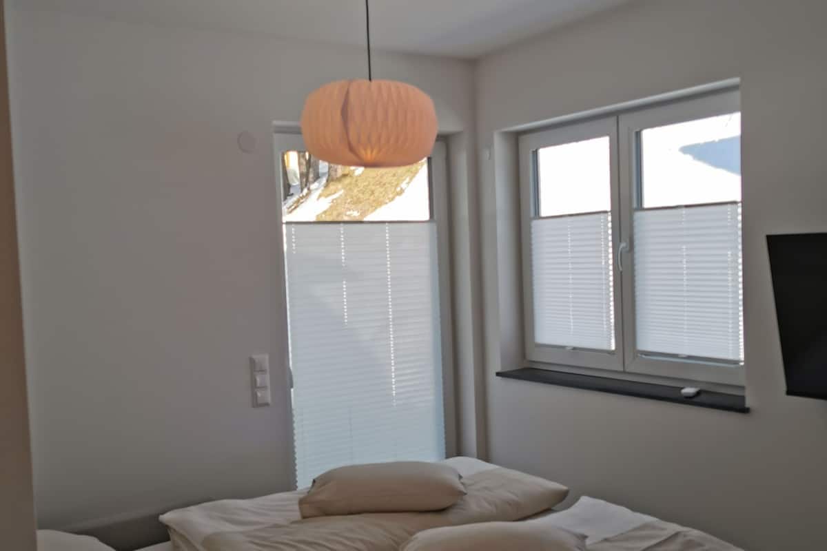 A bedroom with a bed, a hanging light fixture, a window with blinds, and a wall-mounted TV.