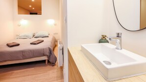 A modern bedroom with a bed, bedside lamps, and a bathroom with a sink and mirror. - Le Balcon de Lavaur – Views & Charm in the Heart of the City (Lavaur)