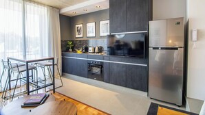 Une cuisine moderne avec des meubles de couleur sombre, un réfrigérateur en acier inoxydable et un four encastré. On y trouve un coin repas avec une table et des chaises, ainsi qu’une fenêtre agrémentée de rideaux légers.