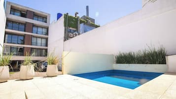 Un área de piscina exterior moderna con piscina azul, plantas en macetas y un edificio al fondo.