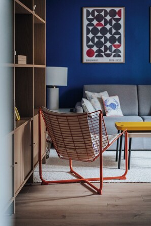 A modern living room with a wooden bookshelf, a red wire chair, a grey sofa, and a yellow table. - Hop Guest Apartment (Meursault)