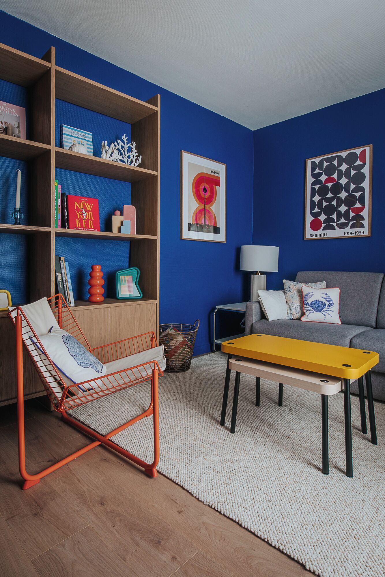 A living room with a blue wall, a wooden bookshelf, a red chair, a yellow table, and a grey sofa.
