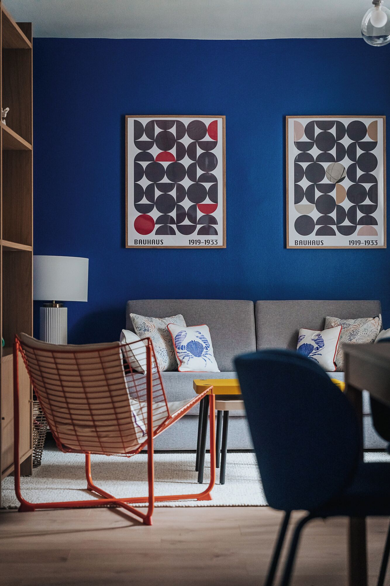 Ein modernes Wohnzimmer mit blauer Akzentwand, zwei gerahmten abstrakten Kunstwerken, einer Couch mit gemusterten Kissen, einem roten Drahtstuhl und einem kleinen Tisch mit einer Schachtel.