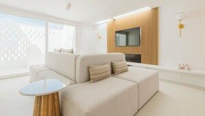 Ein modernes Wohnzimmer mit einer Eckcouch, einem kleinen runden Tisch, einem Flachbildfernseher an einer Wand aus Holzpaneelen und einem großen Fenster, das natürliches Licht hereinlässt.