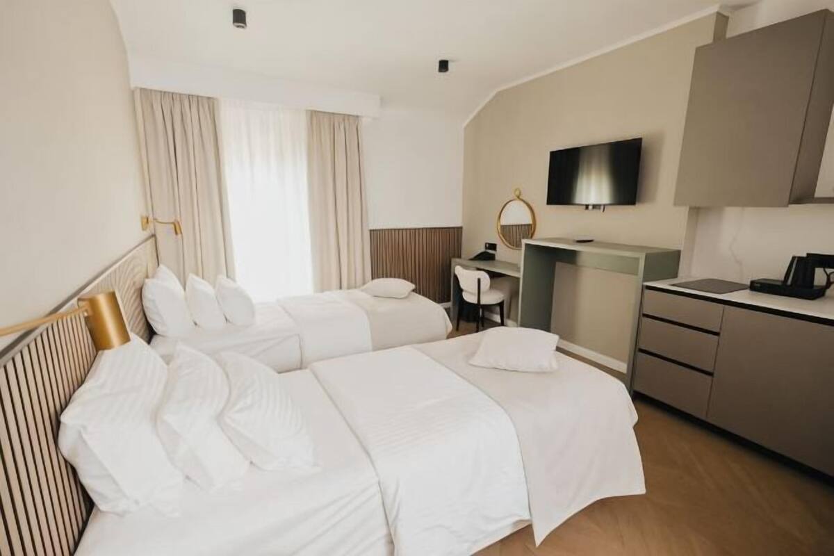 A hotel room with two beds, a desk, a TV, and a kitchenette.