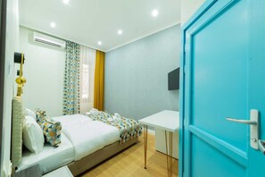 A hotel room with a bed, a desk, a blue door, and an air conditioning unit. - Marita (Tbilisi)