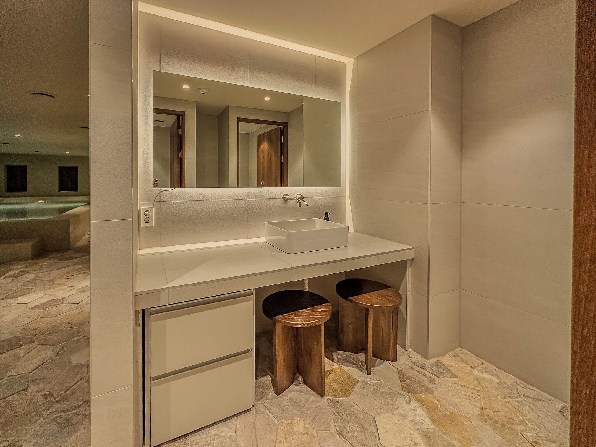 A modern bathroom with a large mirror, a white sink, and two wooden stools.