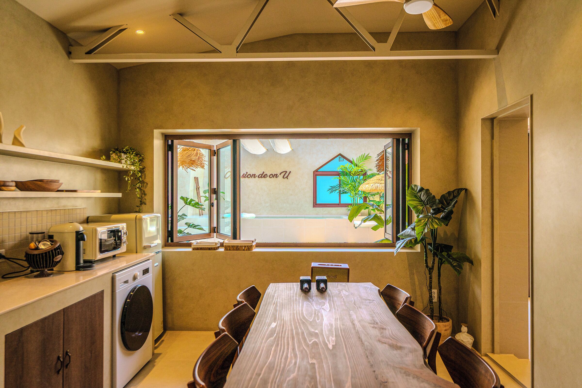 Una cocina compacta con una mesa de madera, electrodomésticos integrados y un ventanal con vista a una casa.