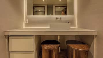 A bathroom with a white sink, a mirror above it, and a small refrigerator below. There are two wooden stools in front of the sink.