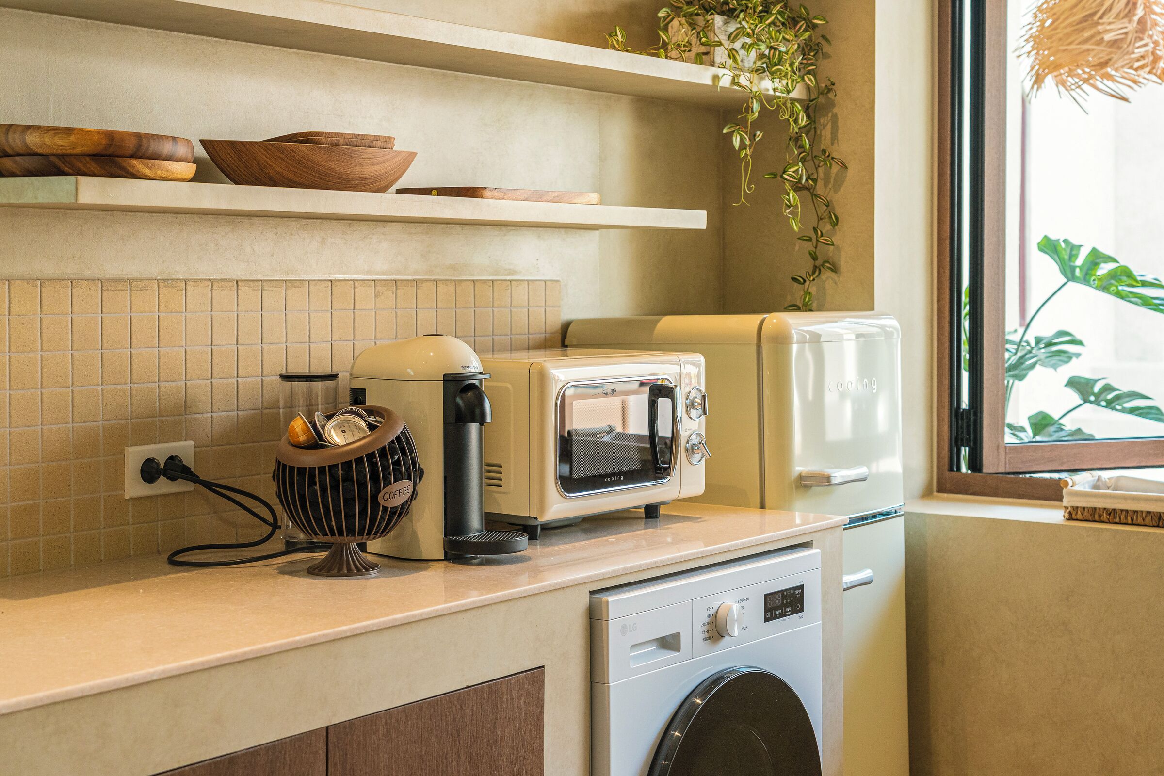 Una cocina con tostadora, cafetera, lavadora y ventana con planta.