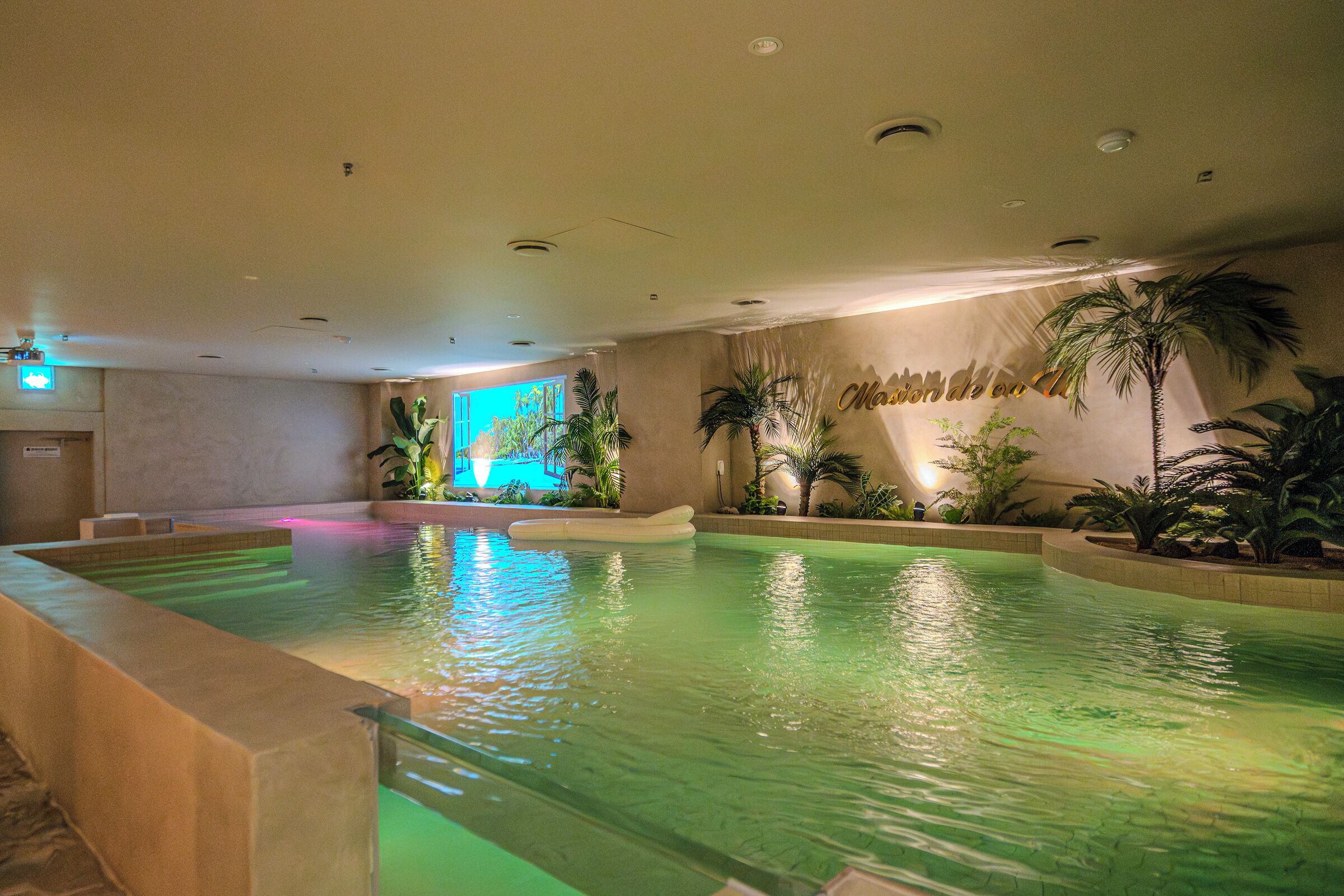 An indoor swimming pool with clear water, surrounded by palm plants and a wall with a mural.