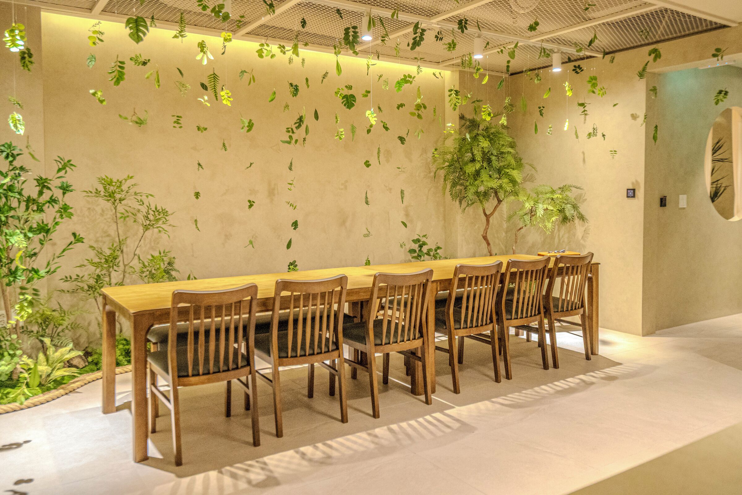 Un comedor con una mesa de madera larga y sillas, rodeado de plantas colgantes y un árbol grande.