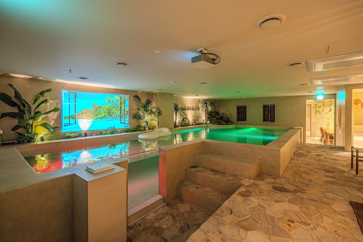 A spacious indoor swimming pool with a stone floor, surrounded by various plants and a large window with a tropical view.