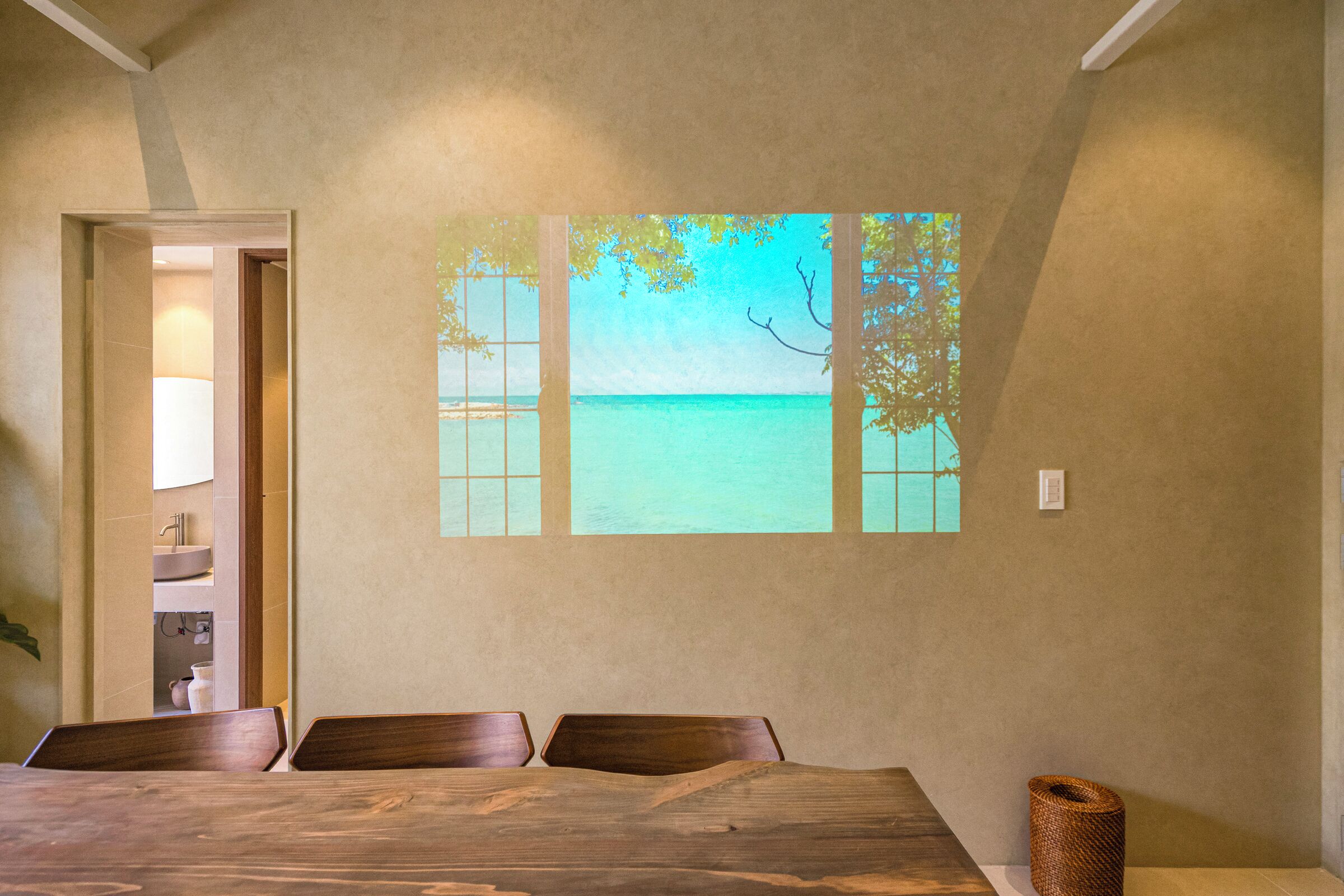 A dining area with a wooden table and chairs, a view of the sea through a window, and a door leading to a bathroom.