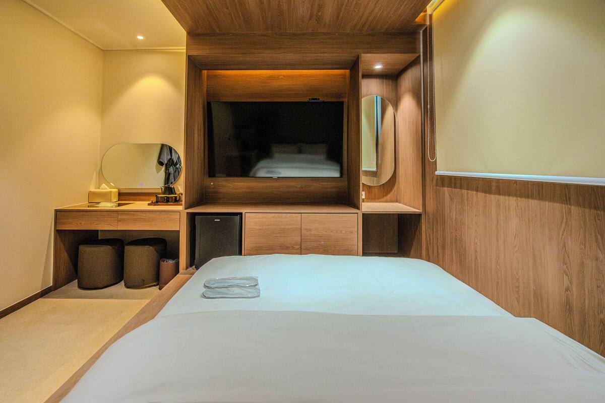 A compact hotel room with a bed, a small desk with a mirror, a nightstand, and a built-in wardrobe.