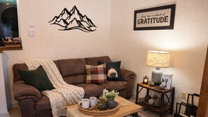 A cozy living room with a brown sofa, a blanket, and a coffee table with mugs and a vase. A framed sign on the wall reads "always have an attitude of GRATITUDE." - Escape to Tranquility at Mountain Top Condo! (Somerset)