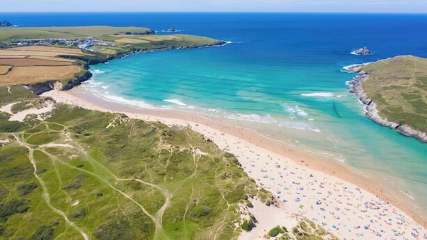 Vista aérea de una zona costera con playa, aguas azules cristalinas y campos verdes.