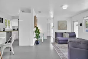 A modern living room with a purple sofa, a dining area with a white table and chairs, and a kitchen with white cabinets and a green countertop.