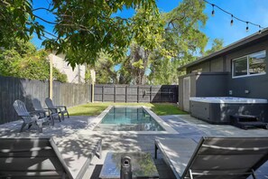 A backyard with a swimming pool, lounge chairs, a hot tub, and a wooden fence.