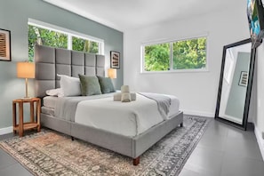 A modern bedroom with a large bed, two bedside lamps, a patterned rug, and a view of greenery through the windows.