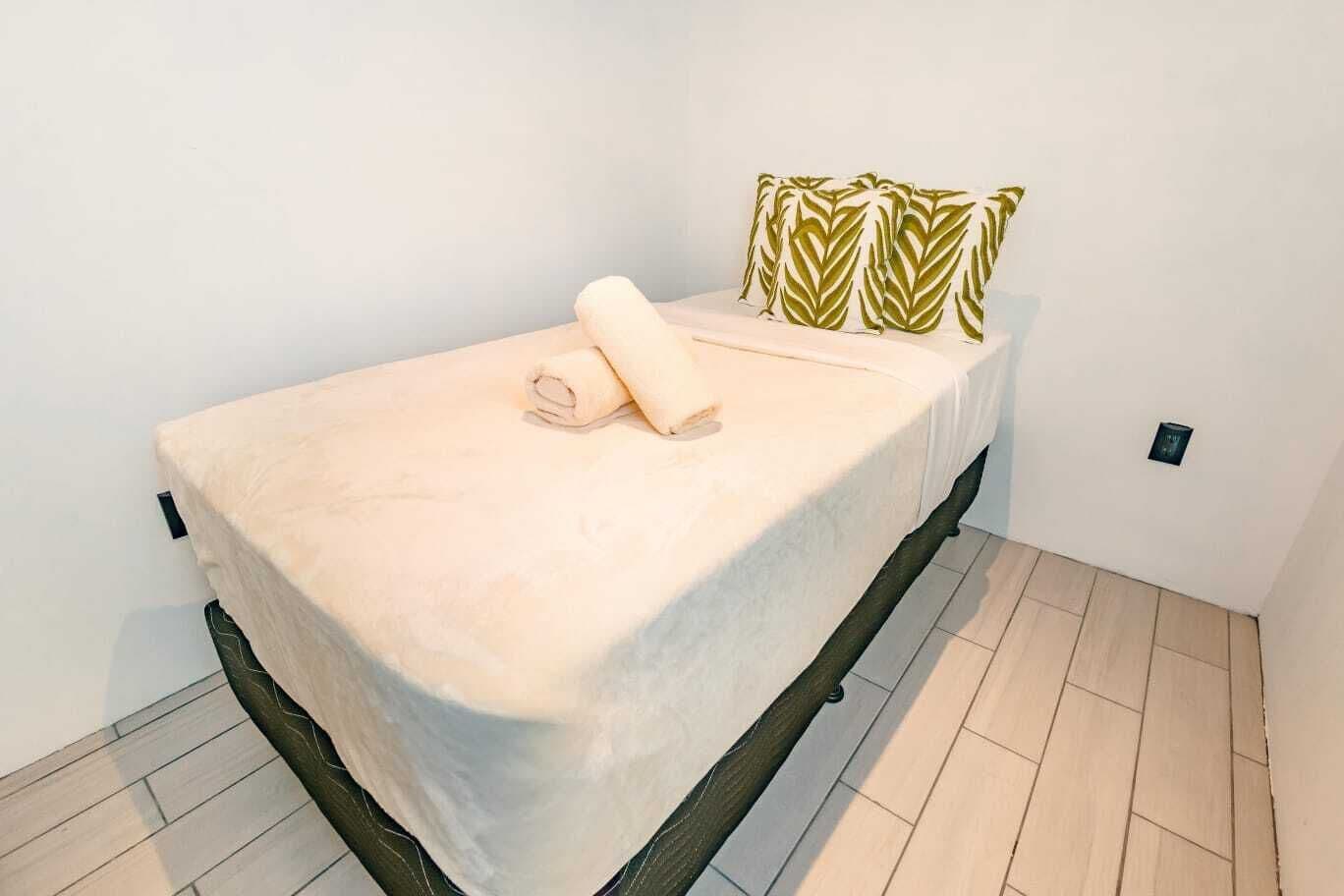 A single bed with a beige cover, a rolled-up towel, and a patterned pillow in a room with white walls and wooden flooring.