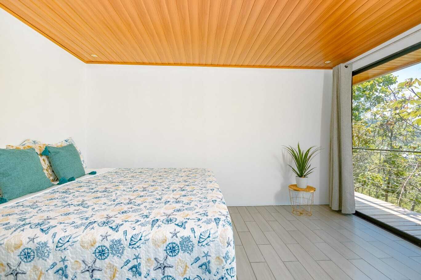 A bedroom with a large bed, a potted plant, and a view of trees through a window.