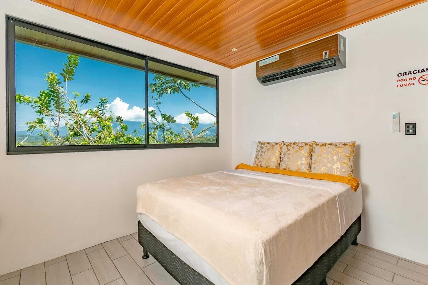 A bedroom with a bed, a window with a view of greenery and mountains, a wooden ceiling, and an air conditioning unit.