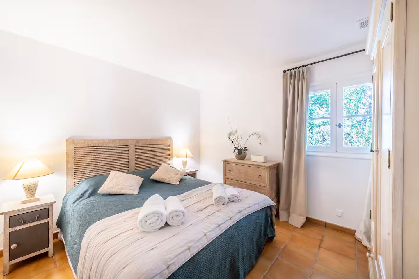 Une chambre à coucher comprenant un lit, des tables de chevet, une commode, une fenêtre avec des rideaux et une vue sur la verdure à l’extérieur.