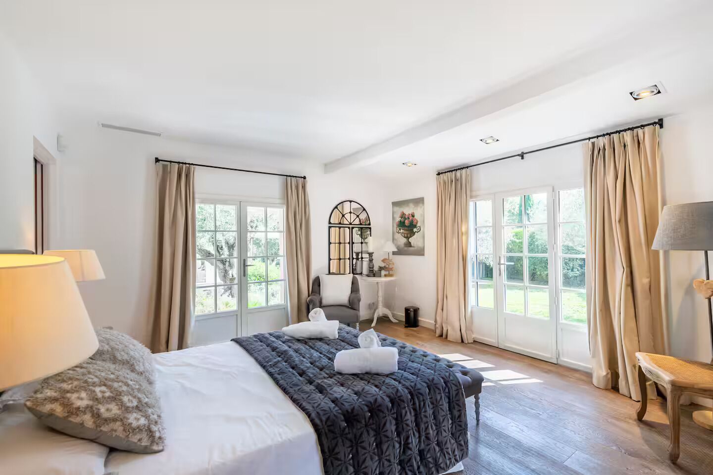 Een ruime slaapkamer met een groot bed, twee ramen met gordijnen, een tafeltje met een lamp en een stoel.