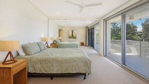 A bedroom with a large bed, two bedside tables with lamps, a ceiling fan, and a sliding glass door leading to a balcony. - Hume Parade 19, Unit 1, Currimundi (Currimundi)