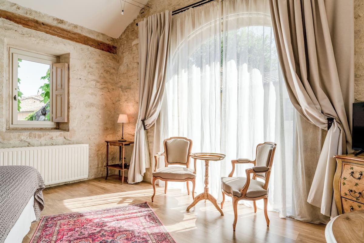Een kamer met een bed, twee stoelen, een tafeltje, een lamp, een radiator en een raam met gordijnen.