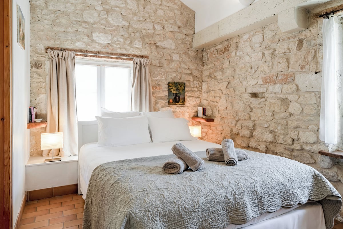Een slaapkamer met een stenen muur, een bed met witte lakens, een raam met gordijnen en een nachtkastje met een lamp.