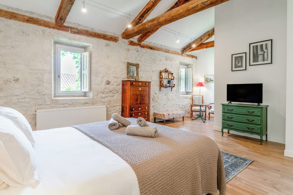 Een slaapkamer met een bed, houten balken, een dressoir, een televisie en een raam.