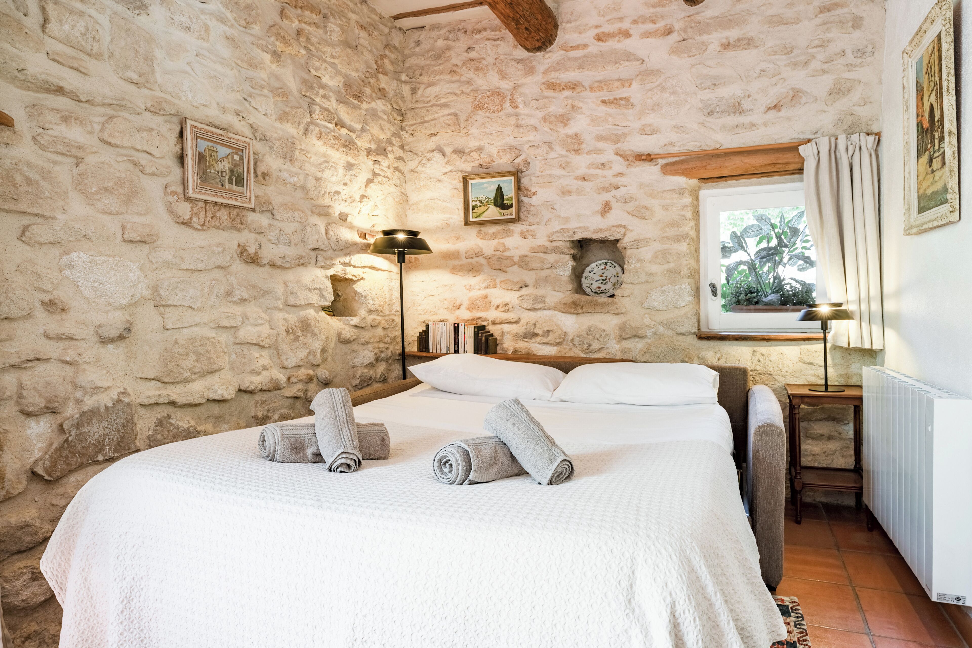 Dormitorio con paredes de piedra, una cama, toallas, una mesita de noche, una lámpara y una ventana con cortinas.