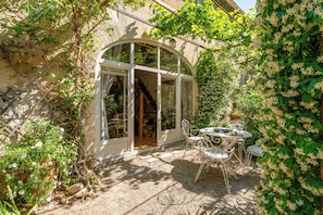 A sunlit patio with a glass door leading to a room, surrounded by greenery and a table with chairs. - Le Clos des Chartreux (Villeneuve-lès-Avignon)